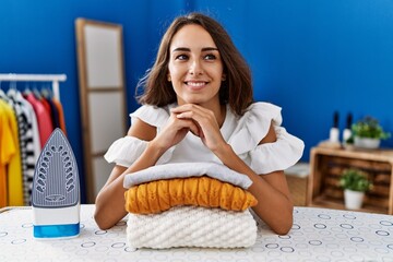 Young hispanic woman smiling confident leaning on folded clothes at laundry room