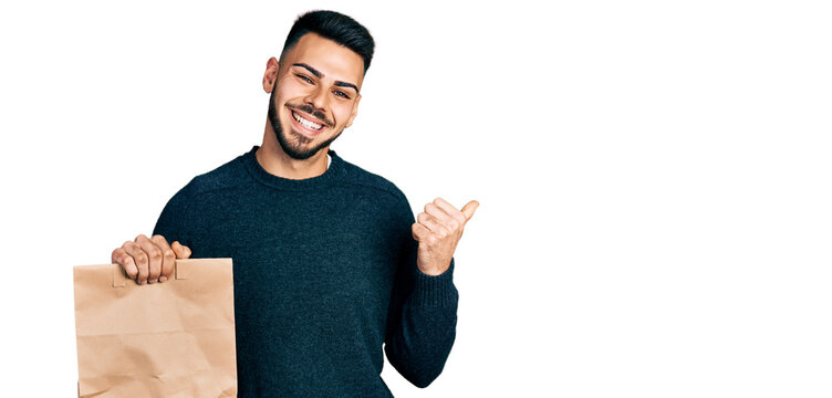 Young hispanic man with beard holding take away paper bag pointing thumb up to the side smiling happy with open mouth