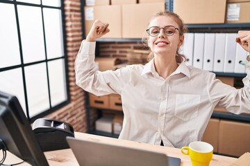 Young caucasian woman working at small business ecommerce using laptop showing arms muscles smiling...