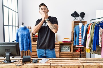 Young hispanic man working at retail boutique laughing and embarrassed giggle covering mouth with hands, gossip and scandal concept