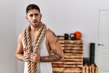 Young hispanic man training with battle rope at the gym in shock face, looking skeptical and...