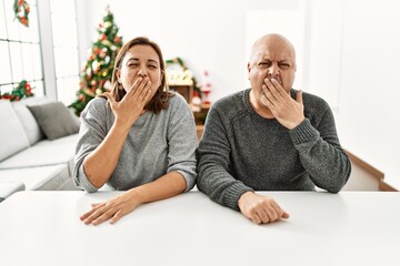 Middle age hispanic couple sitting on the table by christmas tree bored yawning tired covering mouth with hand. restless and sleepiness.