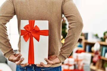 Young hispanic man holding christmas gift on his back at home