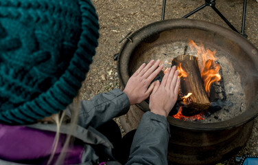 Camping lifestyle concept. Girl wearing beanie hat wariming hands over the campfire on a camping site