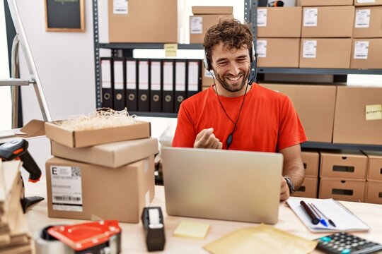 Young hispanic call center agent man working at warehouse beckoning come here gesture with hand inviting welcoming happy and smiling