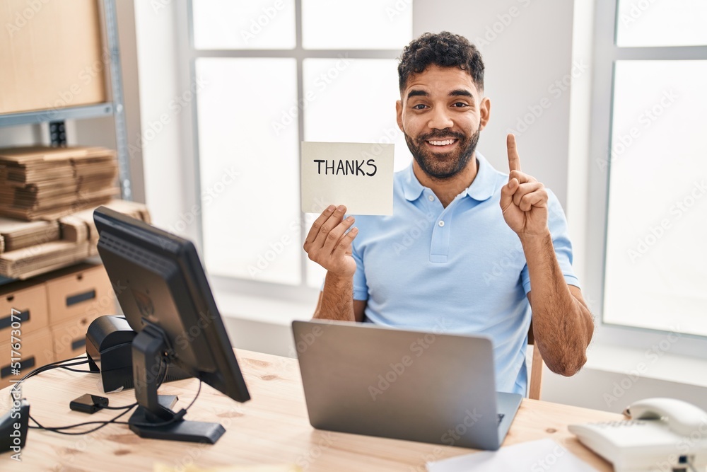 Wall mural Hispanic man with beard working at the office with laptop holding thanks banner smiling with an idea or question pointing finger with happy face, number one