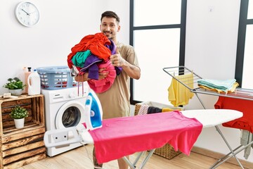 Young handsome man holding laundry ready to iron smiling happy and positive, thumb up doing excellent and approval sign