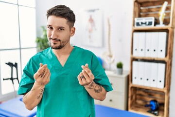 Young physiotherapist man working at pain recovery clinic doing money gesture with hands, asking...