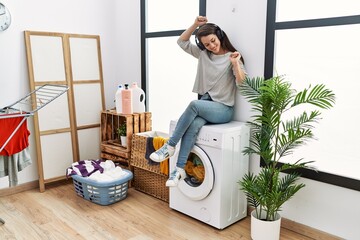 Young hispanic woman listening to music waiting for washing machine at laundry room