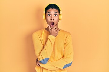 Young african american guy listening to music using headphones looking fascinated with disbelief, surprise and amazed expression with hands on chin