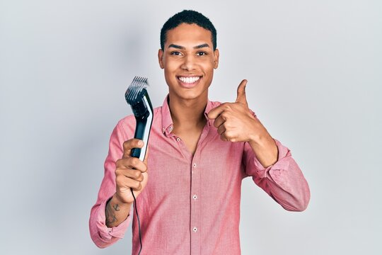 Young african american guy holding electric razor machine smiling happy and positive, thumb up doing excellent and approval sign