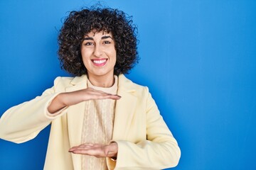 Young brunette woman with curly hair standing over blue background gesturing with hands showing big and large size sign, measure symbol. smiling looking at the camera. measuring concept.