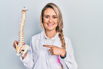Beautiful young blonde doctor woman holding anatomical model of spinal column smiling happy...