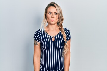 Beautiful young blonde woman wearing casual striped dress puffing cheeks with funny face. mouth inflated with air, crazy expression.