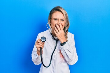 Beautiful young blonde doctor woman holding stethoscope laughing and embarrassed giggle covering...