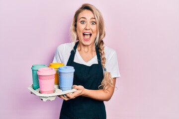 Beautiful young blonde woman wearing waitress apron holding take away cup of coffees celebrating crazy and amazed for success with open eyes screaming excited.