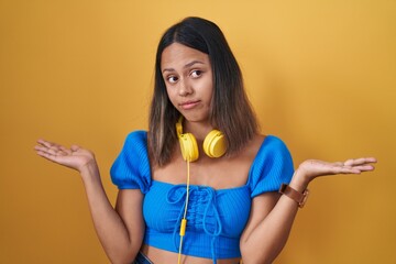 Hispanic young woman standing over yellow background clueless and confused expression with arms and hands raised. doubt concept.
