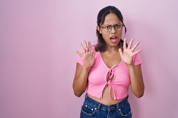 Hispanic young woman standing over pink background wearing glasses afraid and terrified with fear expression stop gesture with hands, shouting in shock. panic concept.