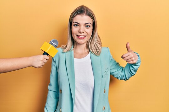 Beautiful caucasian woman being interviewed by reporter holding microphone smiling happy and positive, thumb up doing excellent and approval sign