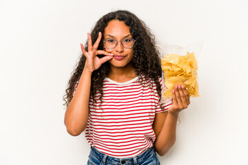 Young hispanic woman holding crips isolated on white background with fingers on lips keeping a...