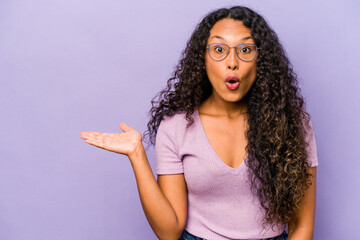 Young hispanic woman isolated on purple background impressed holding copy space on palm.