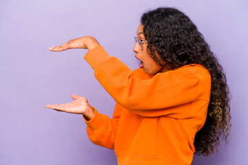 Young hispanic woman isolated on purple background shocked and amazed holding a copy space between hands.