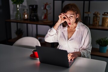 Middle age hispanic woman using laptop at home at night doing ok gesture shocked with surprised face, eye looking through fingers. unbelieving expression.