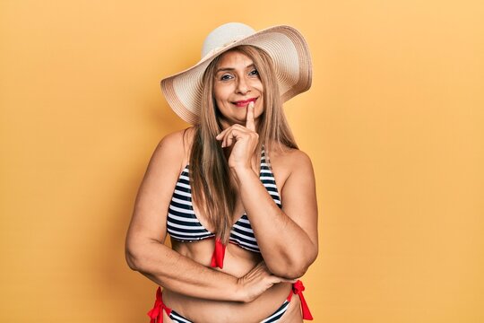 Middle age hispanic woman wearing bikini and summer hat looking confident at the camera with smile with crossed arms and hand raised on chin. thinking positive.