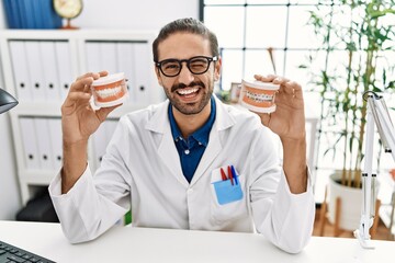 Young hispanic dentist man holding denture with orthodontic braces winking looking at the camera...