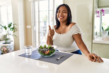 Obraz na płótnie Canvas Young hispanic woman eating healthy salad at home doing happy thumbs up gesture with hand. approving expression looking at the camera showing success.