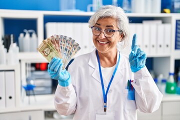 Middle age woman with grey hair working at scientist laboratory holding polish zloty banknotes surprised with an idea or question pointing finger with happy face, number one