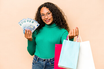 Young hispanic woman shopping and holding bank notes isolated on beige background