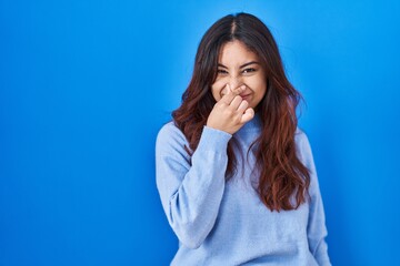 Hispanic young woman standing over blue background smelling something stinky and disgusting, intolerable smell, holding breath with fingers on nose. bad smell