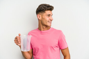 Young caucasian man holding measuring jug isolated on white background looks aside smiling, cheerful and pleasant.