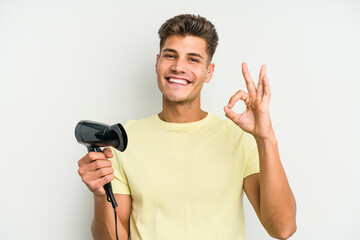 Young caucasian man holding hairdryer isolated on white background cheerful and confident showing ok gesture.