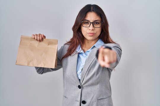 Hispanic young woman holding take away bag pointing with finger to the camera and to you, confident gesture looking serious