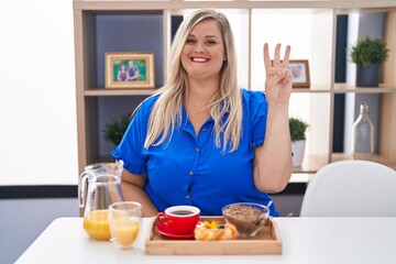 Obraz na płótnie Canvas Caucasian plus size woman eating breakfast at home showing and pointing up with fingers number three while smiling confident and happy.