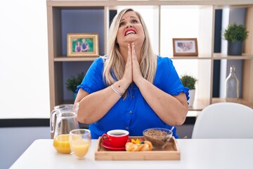 Caucasian plus size woman eating breakfast at home begging and praying with hands together with hope expression on face very emotional and worried. begging.