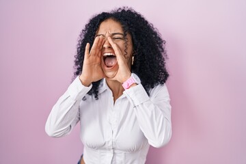 Hispanic woman with curly hair standing over pink background shouting angry out loud with hands...
