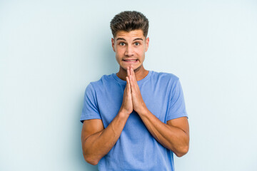 Young caucasian man isolated on blue background holding hands in pray near mouth, feels confident.