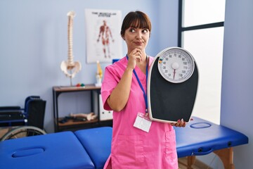 Young brunette woman as nutritionist holding weighing machine serious face thinking about question...