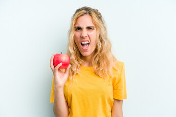 Young caucasian woman holding an apple isolated on blue background screaming very angry and aggressive.