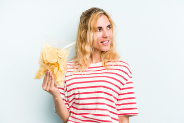 Young caucasian woman holding a bag of chips isolated on blue background looks aside smiling, cheerful and pleasant.