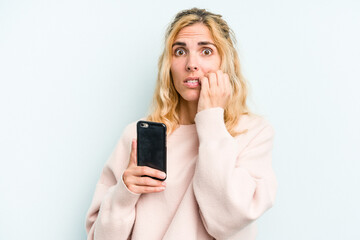 Young caucasian woman holding mobile phone isolated on blue background biting fingernails, nervous...