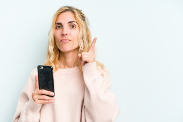 Fototapeta premium Young caucasian woman holding mobile phone isolated on blue background showing number one with finger.