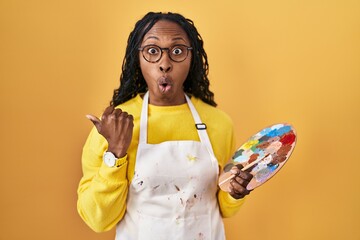 African woman holding painter palette surprised pointing with hand finger to the side, open mouth...