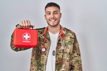 Young hispanic doctor wearing camouflage army uniform holding first aid kit looking positive and...