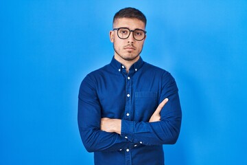 Young hispanic man wearing glasses over blue background skeptic and nervous, disapproving...