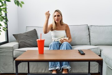 Obraz na płótnie Canvas Young blonde woman eating popcorn sitting on the sofa annoyed and frustrated shouting with anger, yelling crazy with anger and hand raised