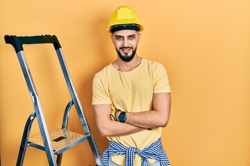 Handsome man with beard by construction stairs wearing hardhat happy face smiling with crossed arms looking at the camera. positive person.